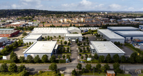 Aerial view of an industrial estate with multiple white-roofed warehouses, surrounded by greenery, with residential buildings in the background.
