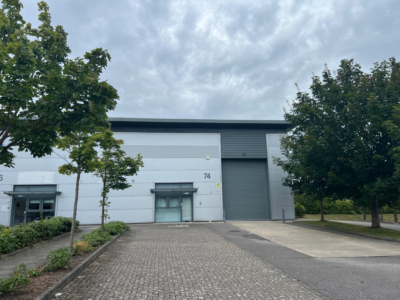 A modern industrial building with a gray exterior, labeled 74, features a large garage door, a smaller entry door, and is surrounded by trees and paving under a cloudy sky.