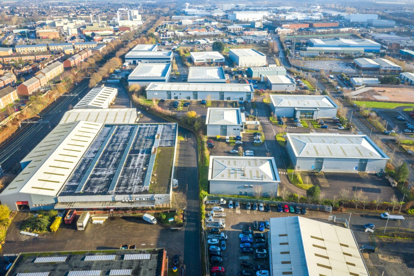 Aerial view of an industrial estate with multiple warehouses, office buildings, parking lots, and adjacent residential areas under daylight.