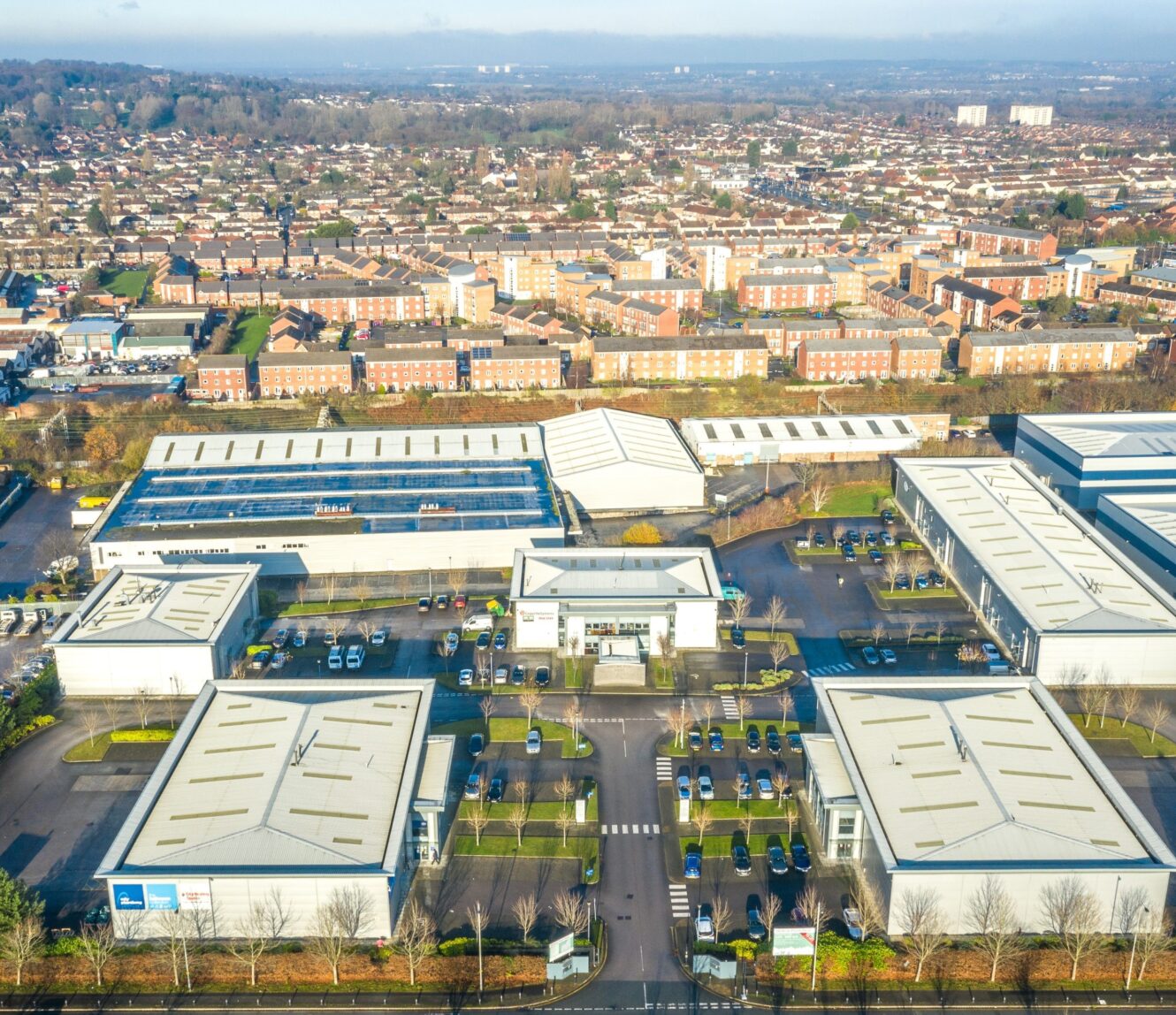 Aerial view of a business park with several modern industrial buildings, parking lots, and a residential neighborhood in the background.
