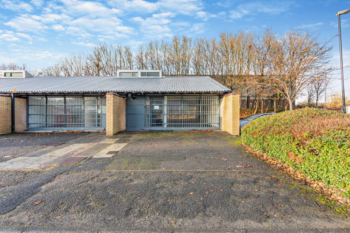 Single-story brick building with two gray metal garage doors, adjacent hedge, and a backdrop of leafless trees under a blue sky.