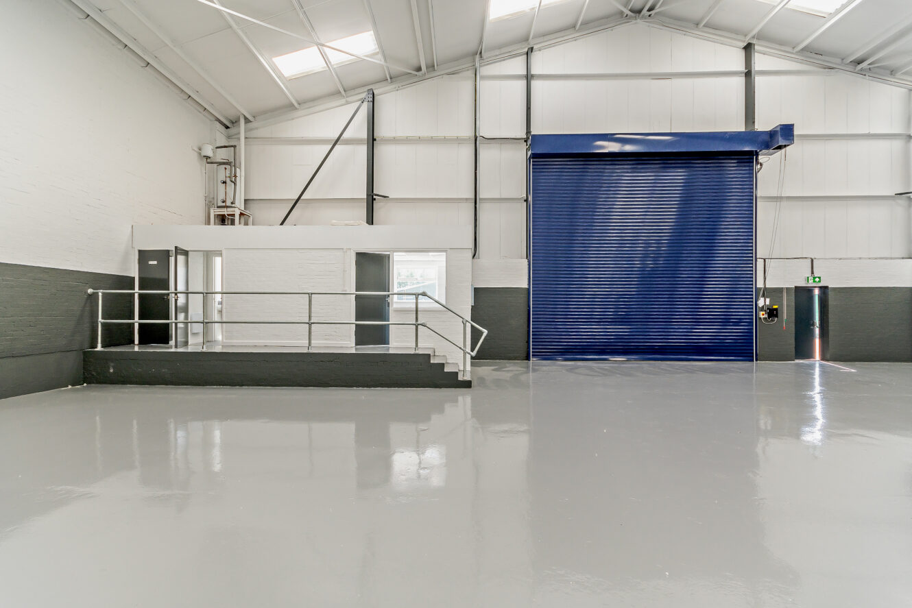 A clean, empty industrial warehouse with a small office area, metal stairs, shiny gray floor, and a large blue roller shutter door.