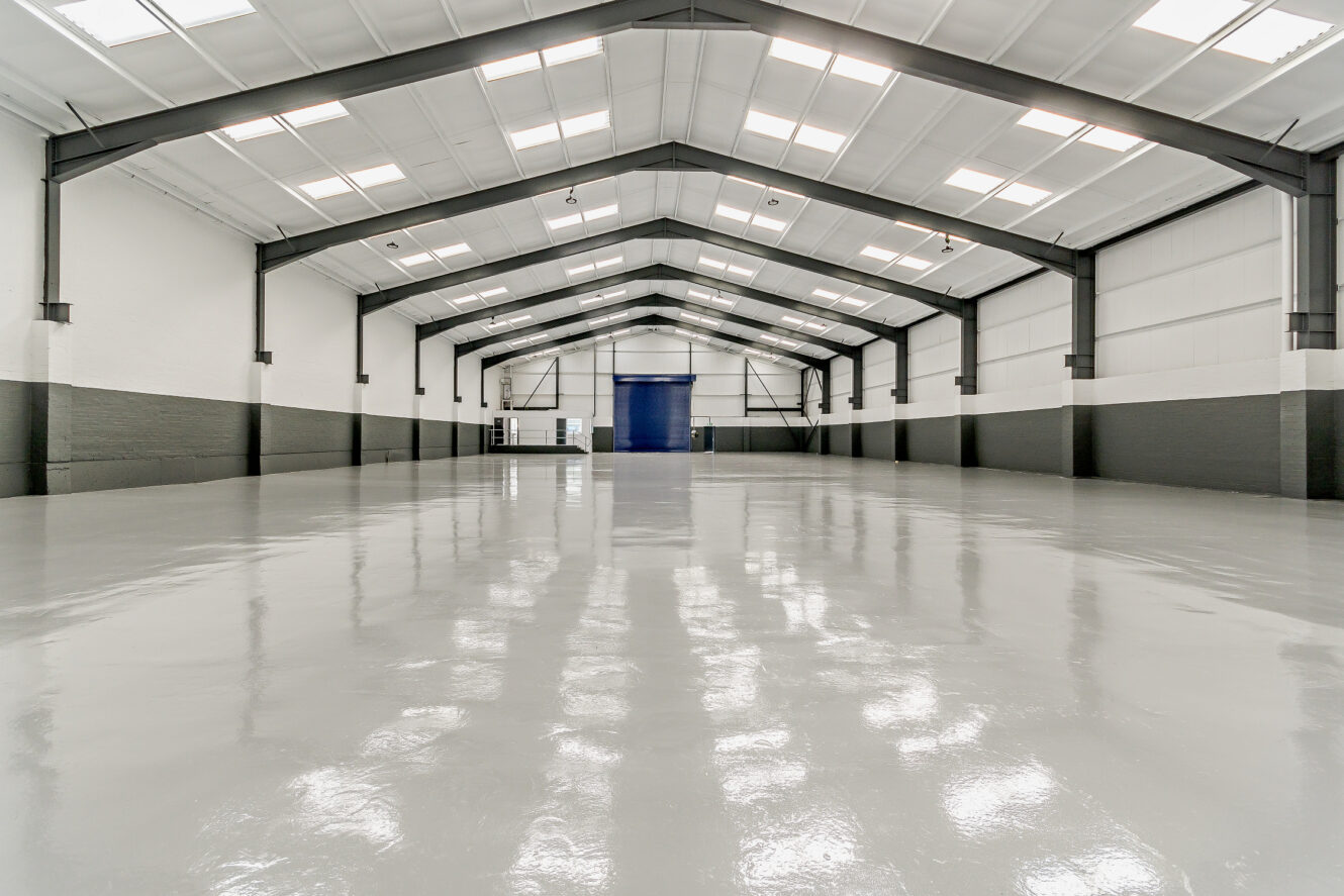 Large empty warehouse interior with high ceilings, white and gray walls, exposed steel beams, and polished concrete floor.