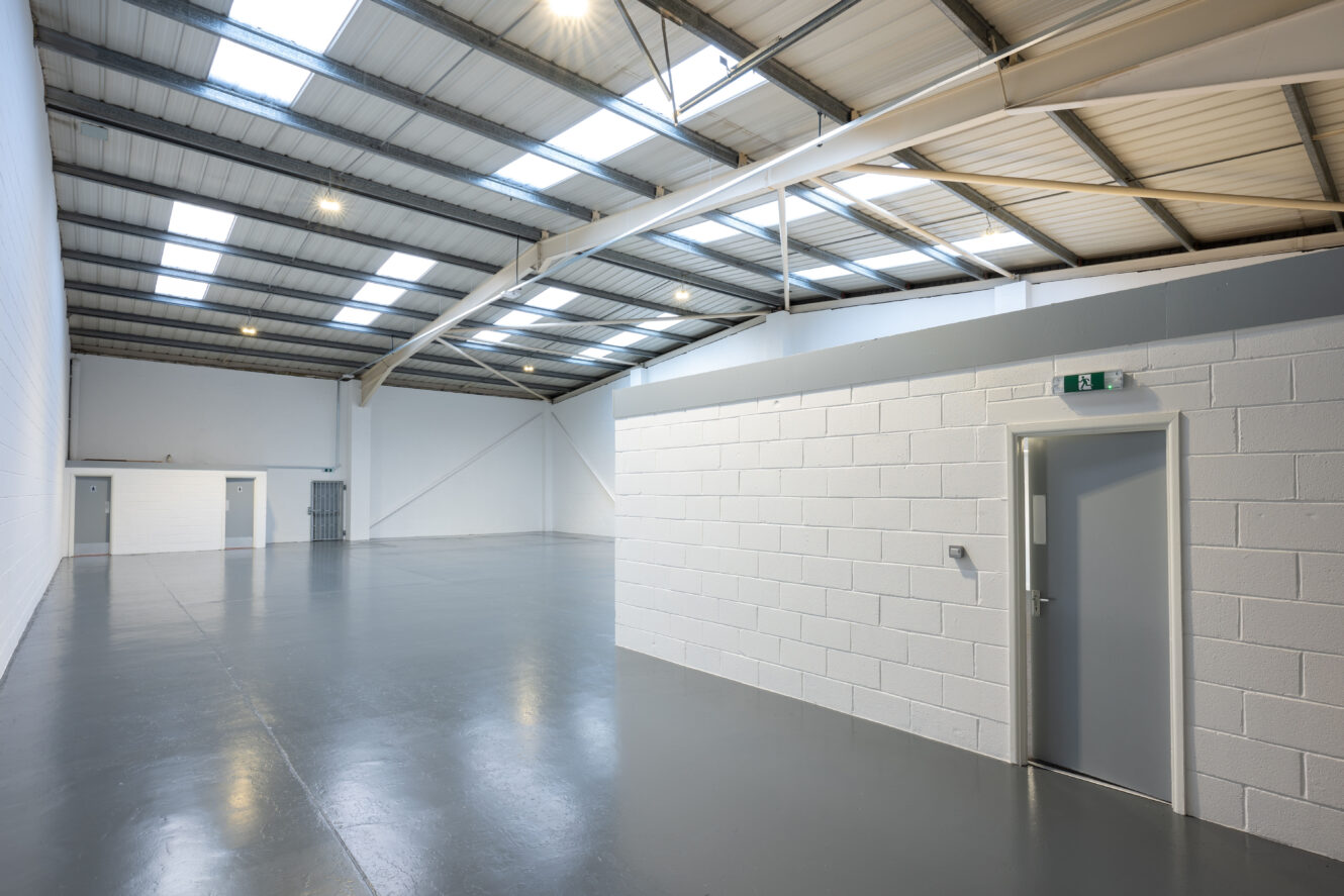 Large, empty industrial warehouse with gray floor, white walls, high ceilings, skylights, and a closed gray exit door on a white partitioned office area.
