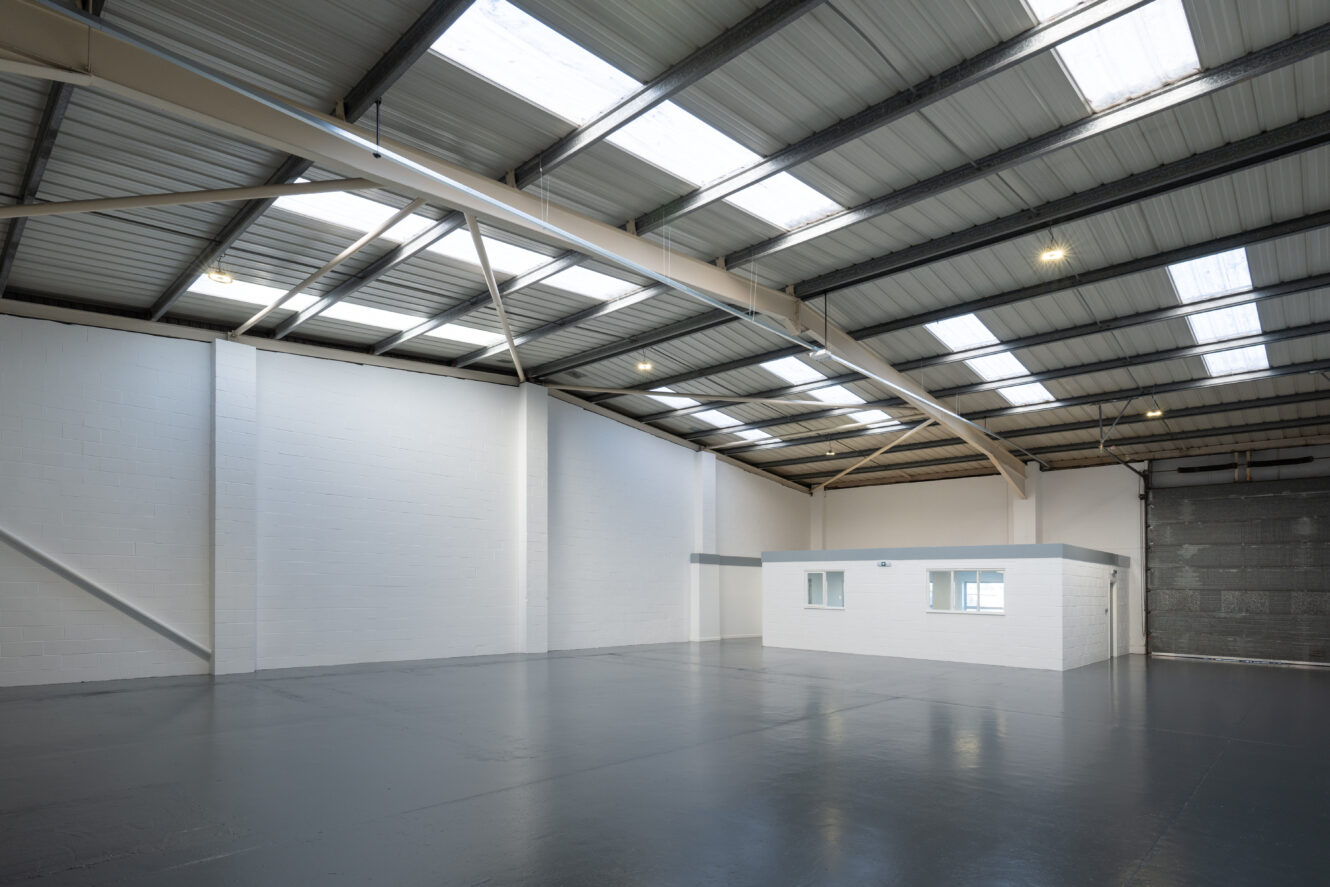 Spacious, empty warehouse with high ceilings, skylights, white walls, grey floor, and a small enclosed office space in one corner.