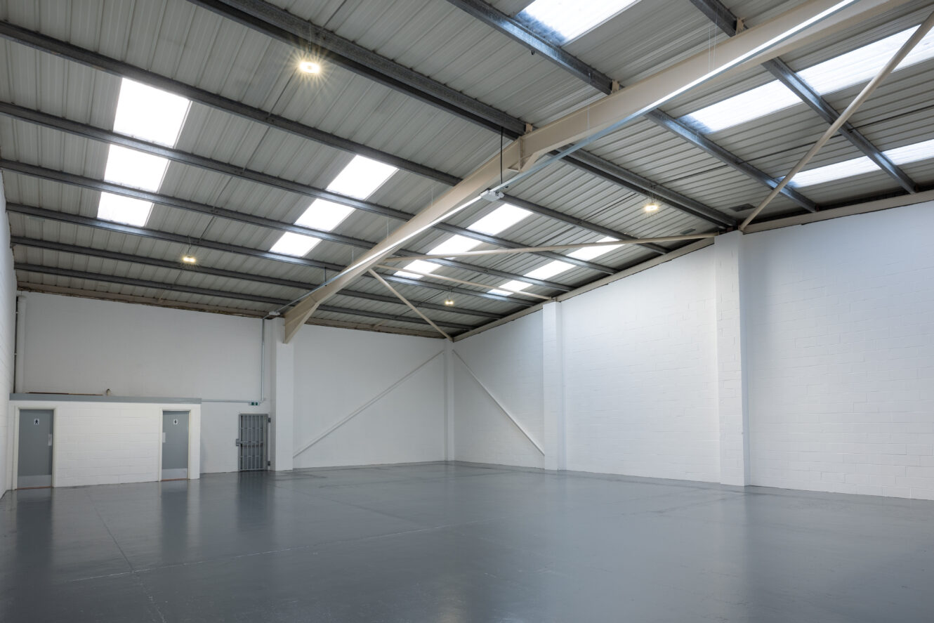 Empty industrial warehouse with gray concrete floor, white walls, metal beams, skylights, and two small doors on the left side.