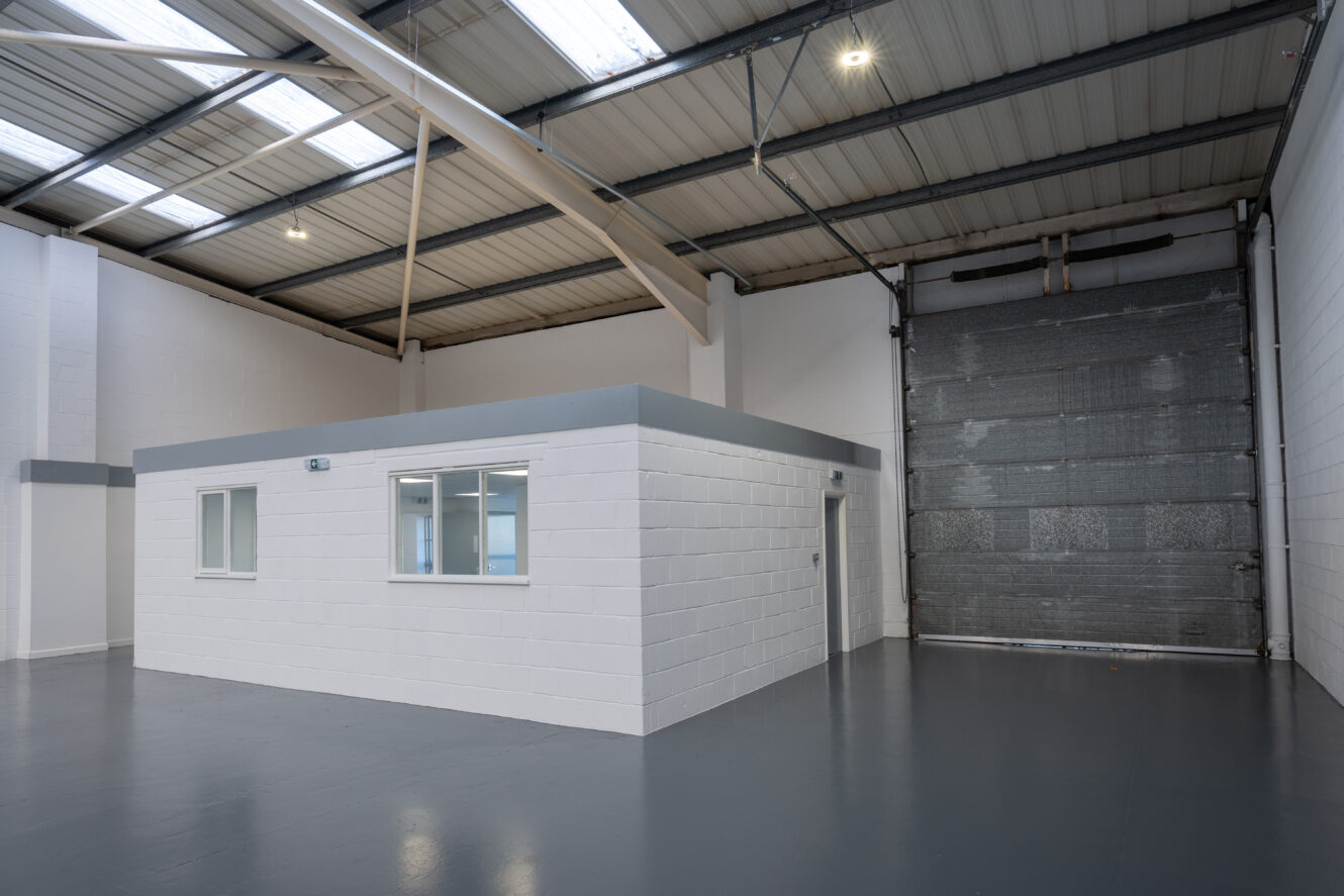 A clean, empty industrial warehouse interior with white walls, grey flooring, a small office structure, and a large roller shutter door.