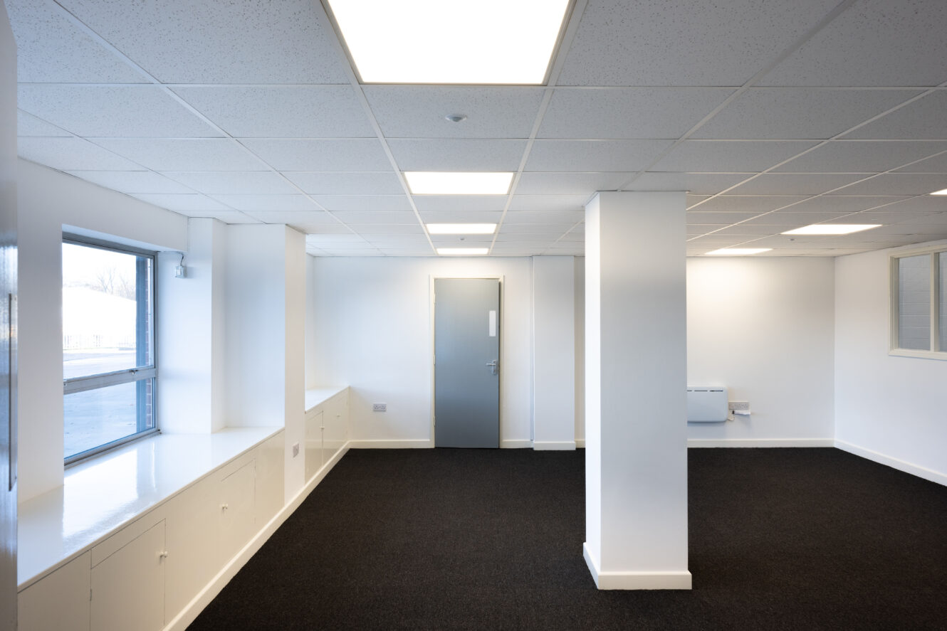 Empty office space with white walls, ceiling lights, black carpet, large window on the left, and a closed gray door at the far end.