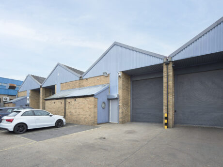 A row of industrial warehouse units with closed roller shutter doors, brick walls, and a white car parked in front on a paved lot.