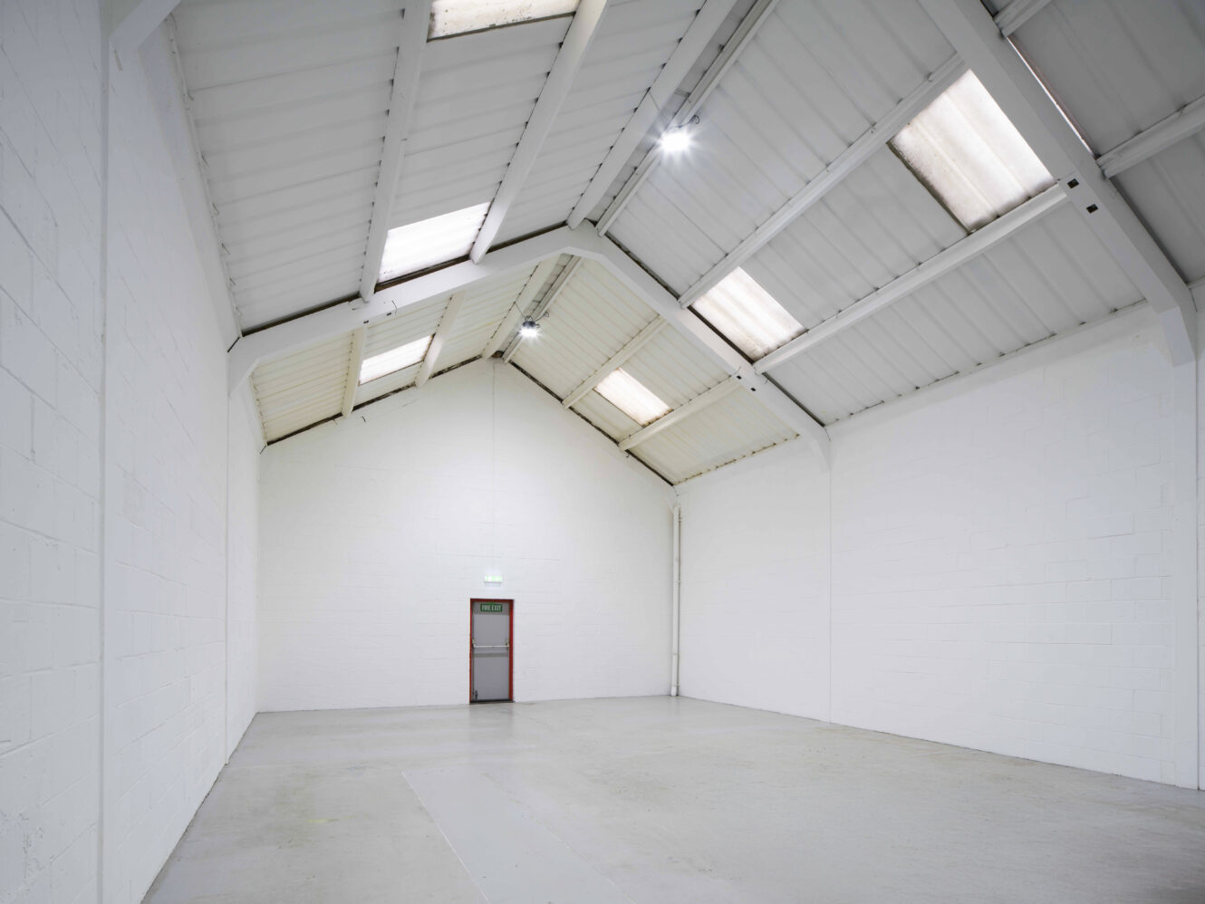 Empty warehouse with white walls, a concrete floor, high ceilings, skylights, and a single closed door at the far end.