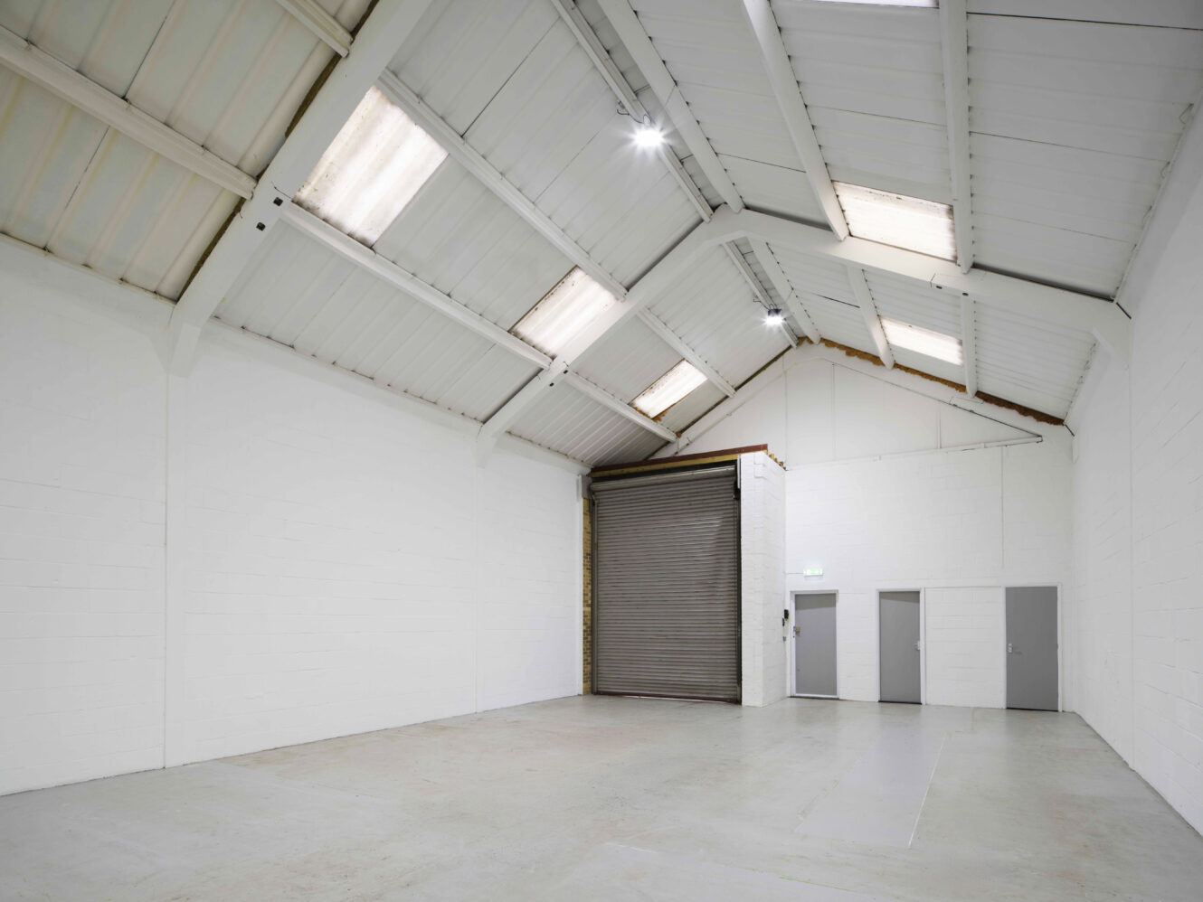 Interior of an empty industrial warehouse with white walls, a concrete floor, high angled ceiling, skylights, a large metal roll-up door, and three gray doors on the right wall.