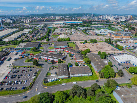 Aerial view of an industrial estate with warehouses, parking lots, and roads, surrounded by green areas and city buildings under a partly cloudy sky.