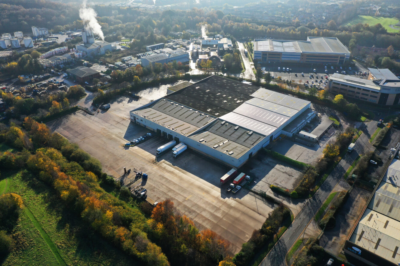 Aerial view of a large warehouse with multiple trucks parked at loading docks, surrounded by industrial buildings, roads, and trees.