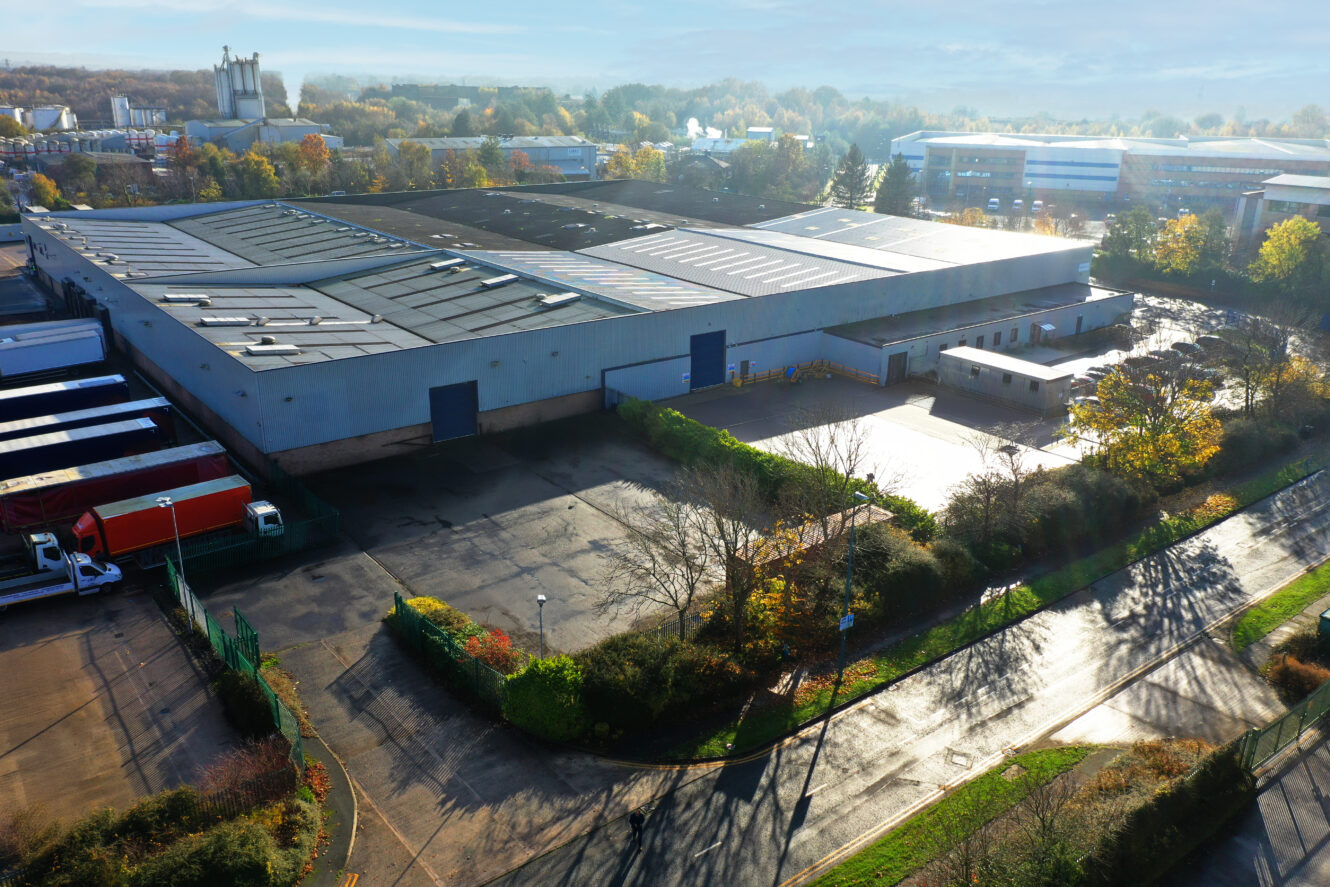 Aerial view of a large industrial warehouse with loading docks, parked trucks, and surrounding greenery on a sunny day.