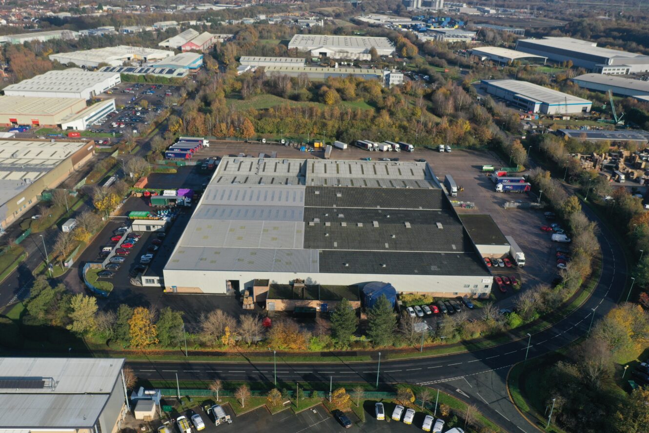 Aerial view of a large industrial warehouse surrounded by parked vehicles, trees, and nearby buildings in an industrial estate.