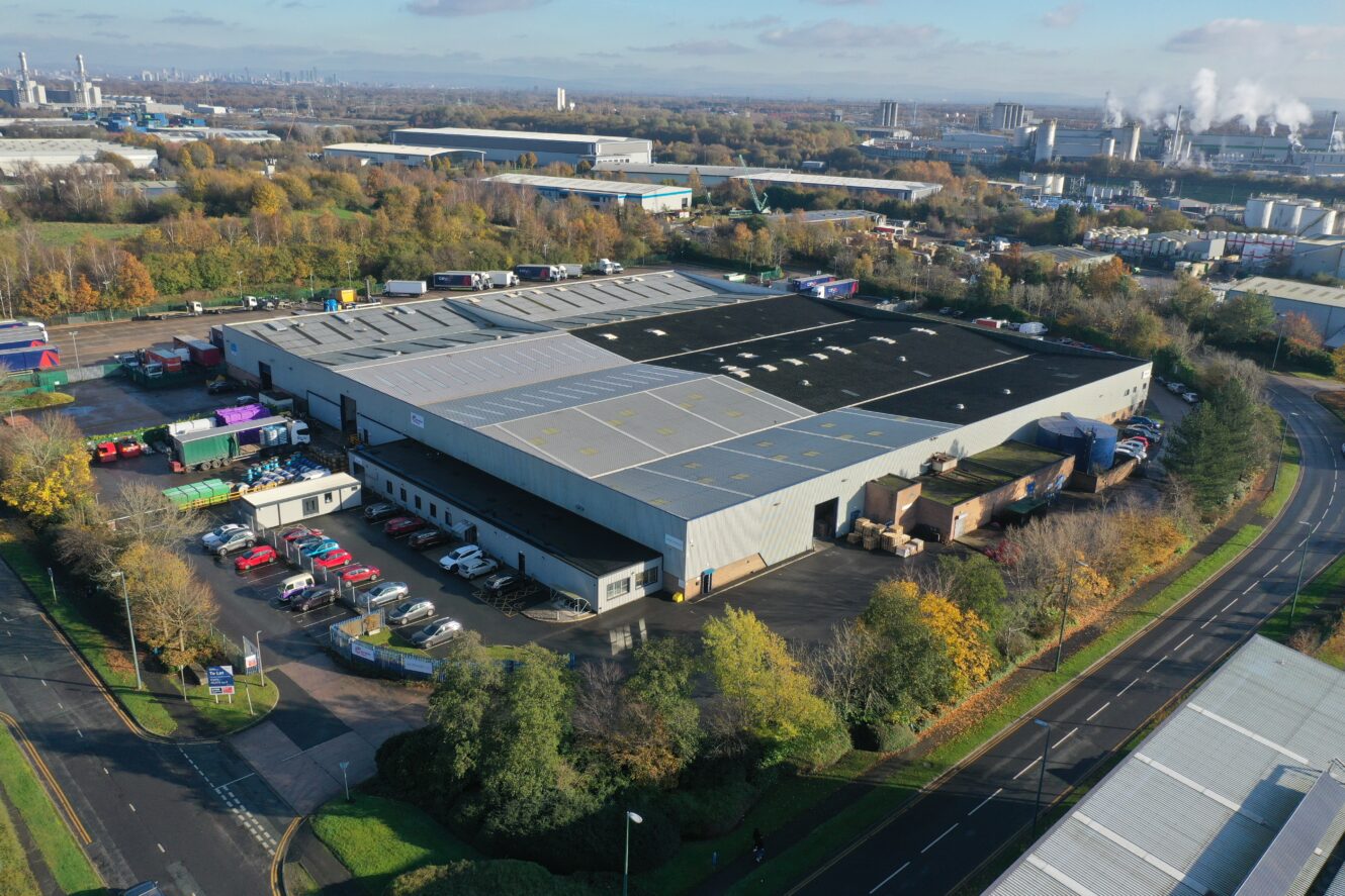 Aerial view of a large industrial warehouse with trucks parked outside, surrounded by trees, roads, and other industrial buildings.