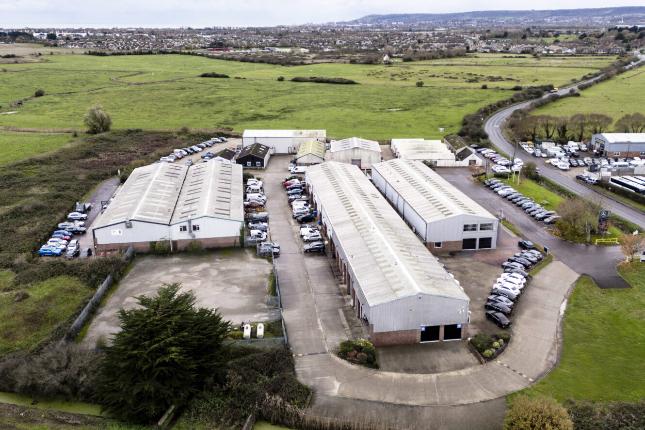 Aerial view of an industrial complex with several large warehouse buildings, parking areas, and surrounding fields, located near a curved road and open green space.