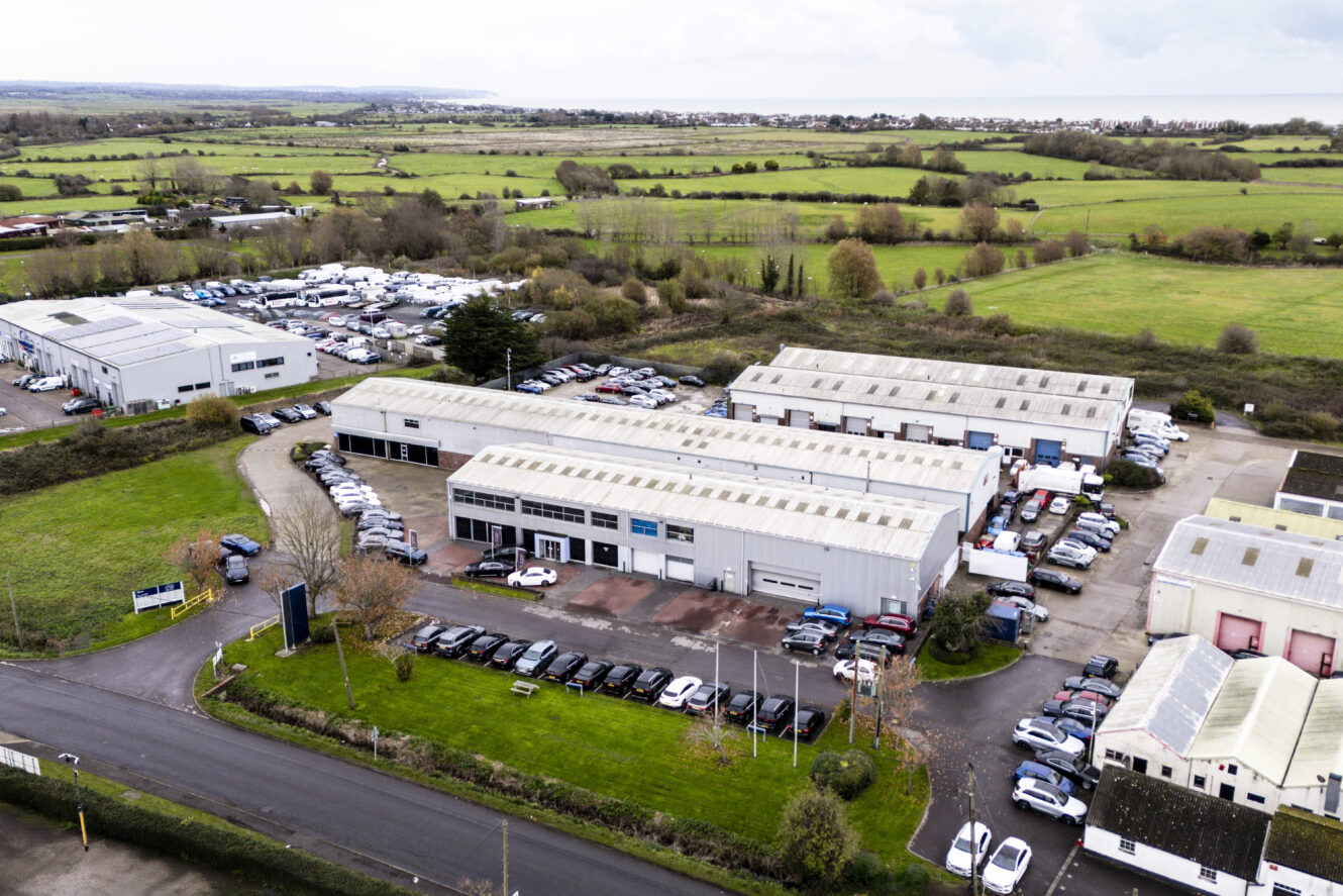 Aerial view of an industrial park with multiple warehouse buildings, surrounding parking lots filled with cars, and green fields in the background.