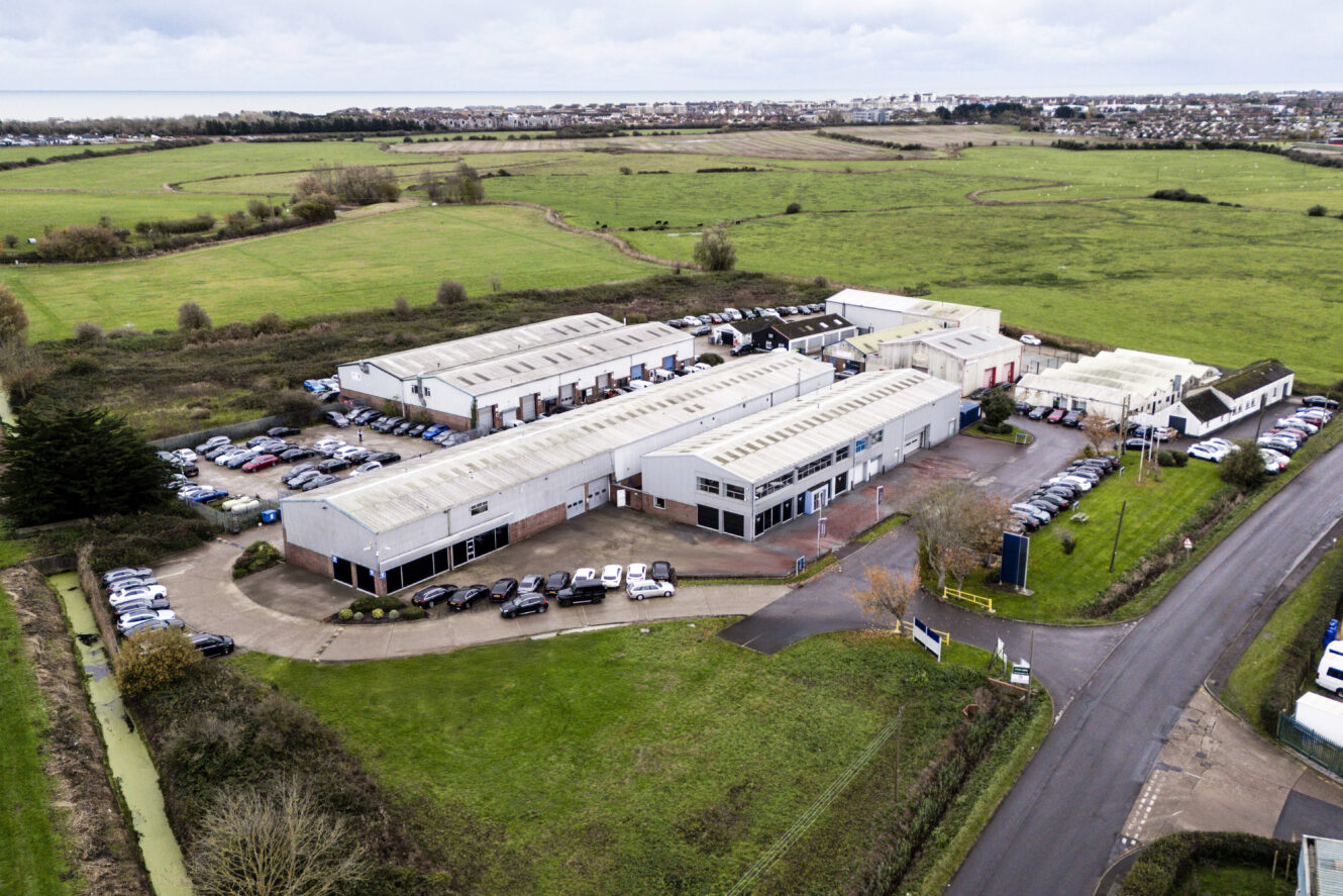 Aerial view of a large industrial building complex with multiple warehouses, surrounded by parked cars, green fields, and a nearby road.
