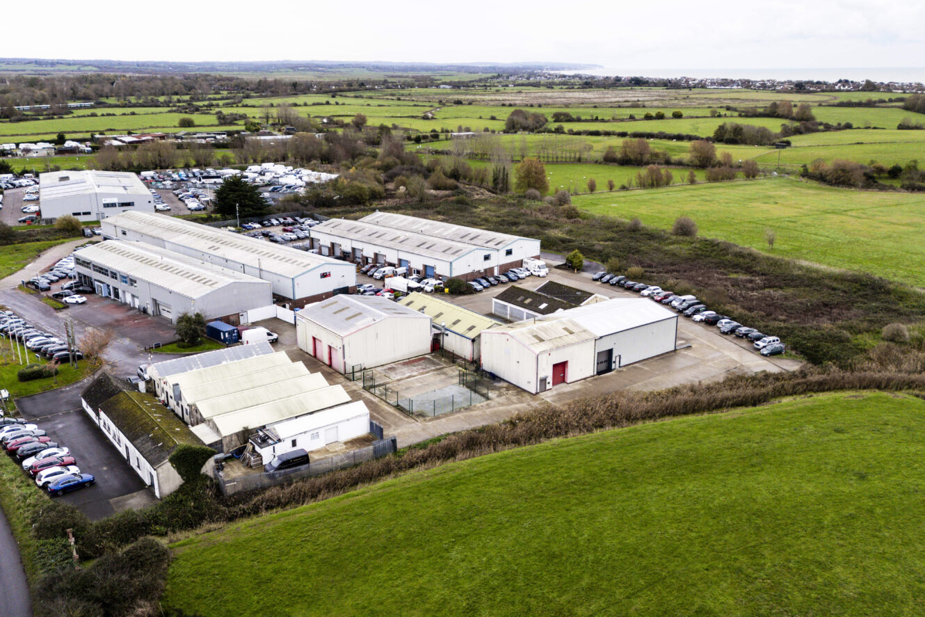 Aerial view of a light industrial complex with multiple warehouses and parked cars, surrounded by green fields and rural landscape.