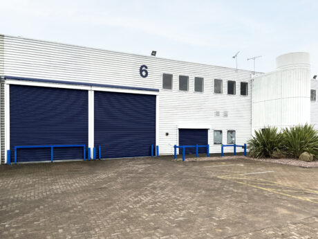 Industrial building with blue roller doors, marked as number 6, surrounded by concrete pavement and a few green plants.