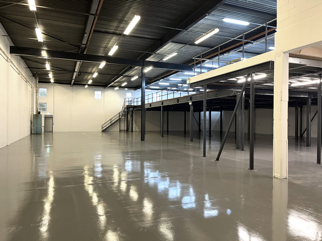 Spacious, empty warehouse interior with polished concrete floors, high ceilings, exposed beams, and a metal mezzanine accessible by stairs.
