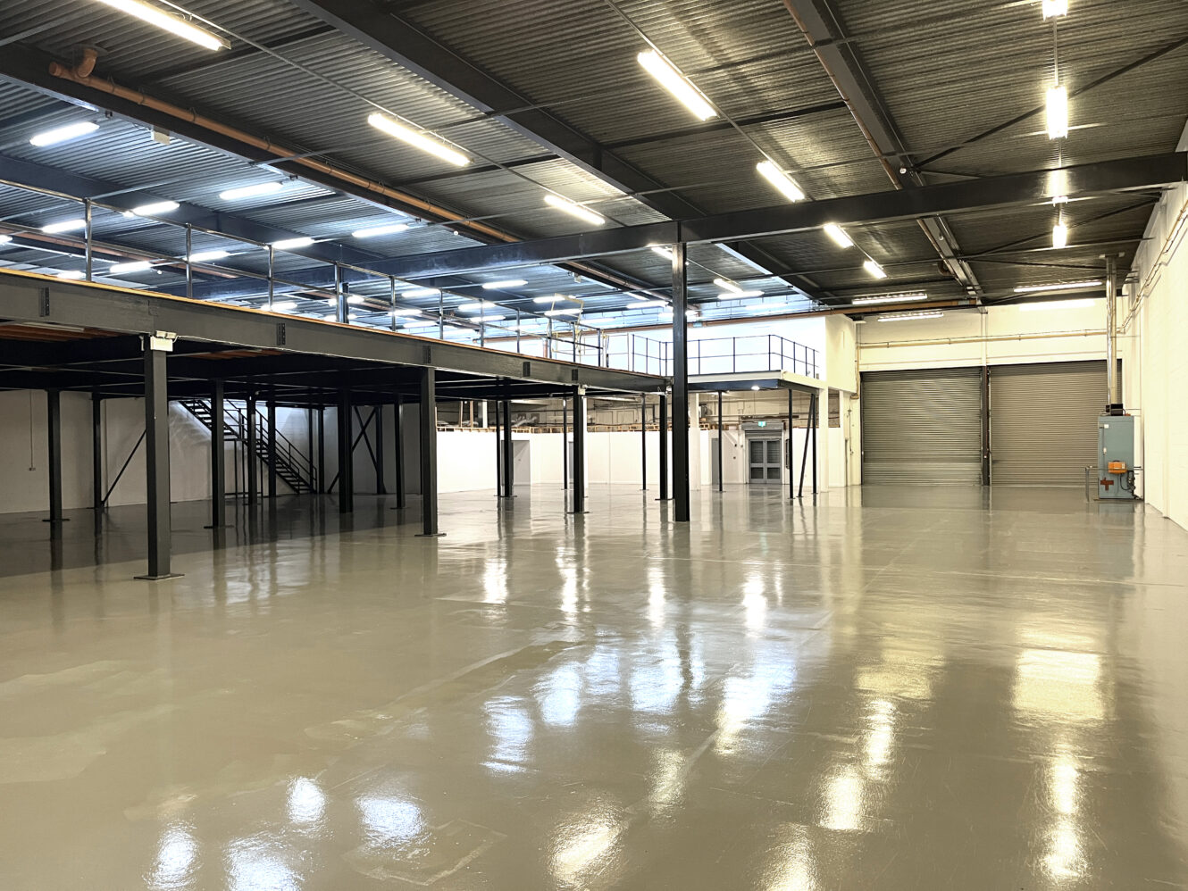 Interior of a large, empty warehouse with polished concrete floors, high ceilings, metal beams, a mezzanine level, and bright overhead lighting.