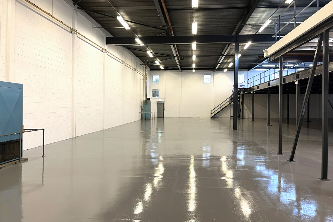 Interior of an empty industrial warehouse with polished concrete floors, white brick walls, overhead lights, and a mezzanine level with metal stairs.
