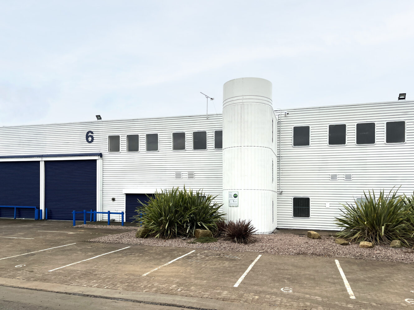 Industrial building with white metal siding, blue doors, number 6 on the wall, small landscaped area with plants, and empty parking spaces in front.