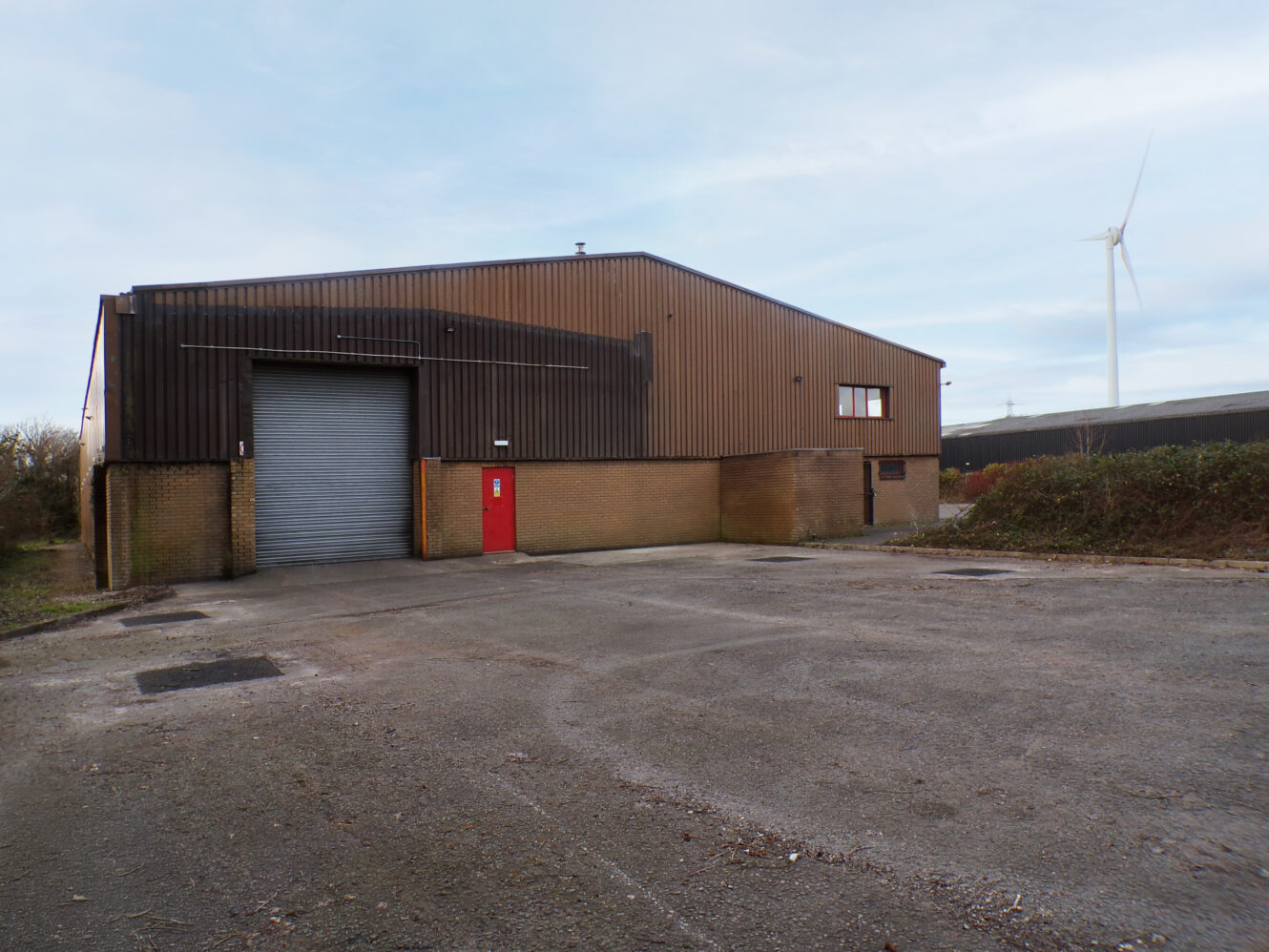 A large brown industrial warehouse with a closed gray roller door and a red side door, set on a paved lot with a wind turbine visible in the background.