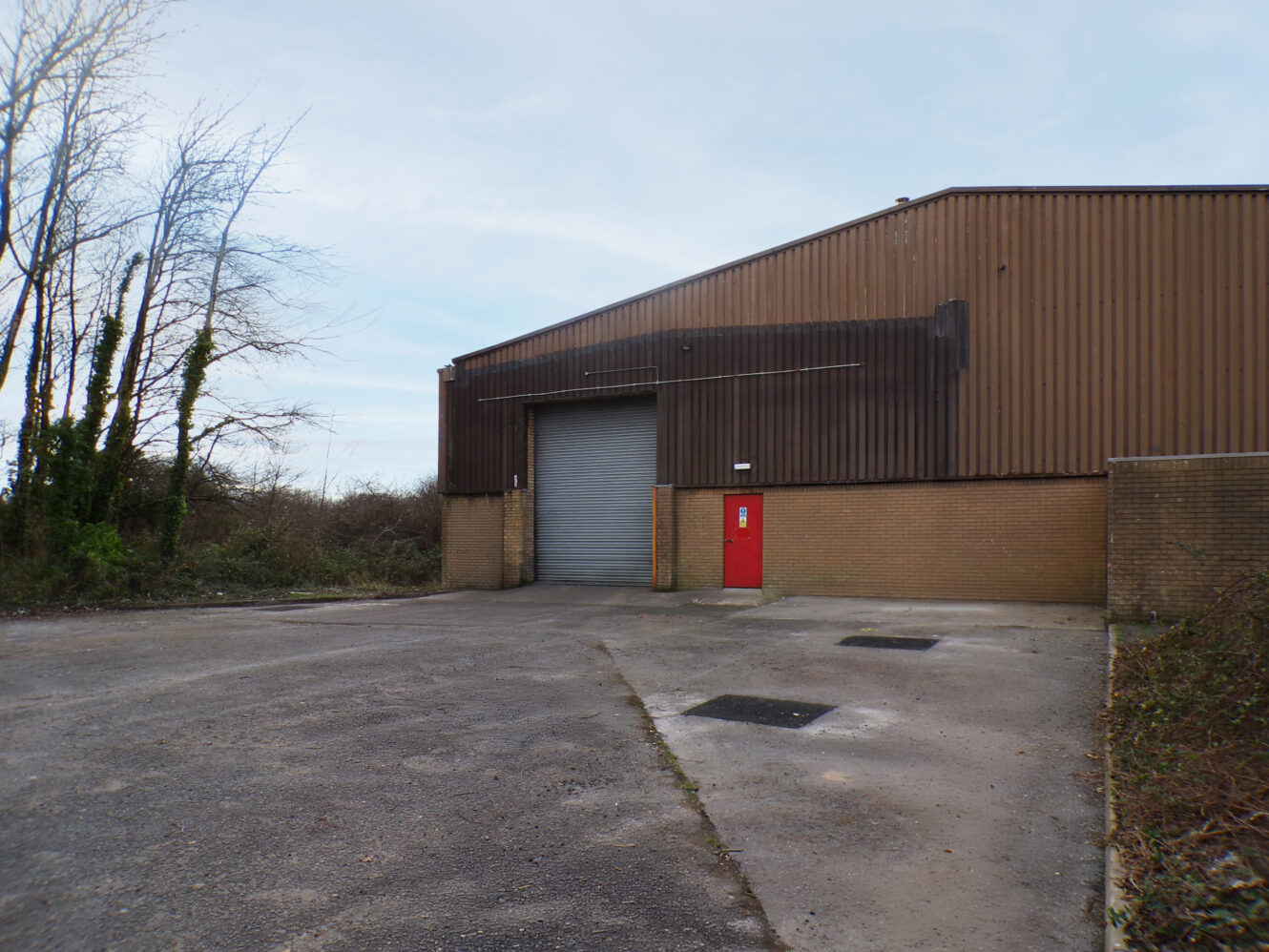 A brown industrial warehouse with a closed metal roller door and a red entrance door, with an empty concrete yard and some overgrown vegetation nearby.