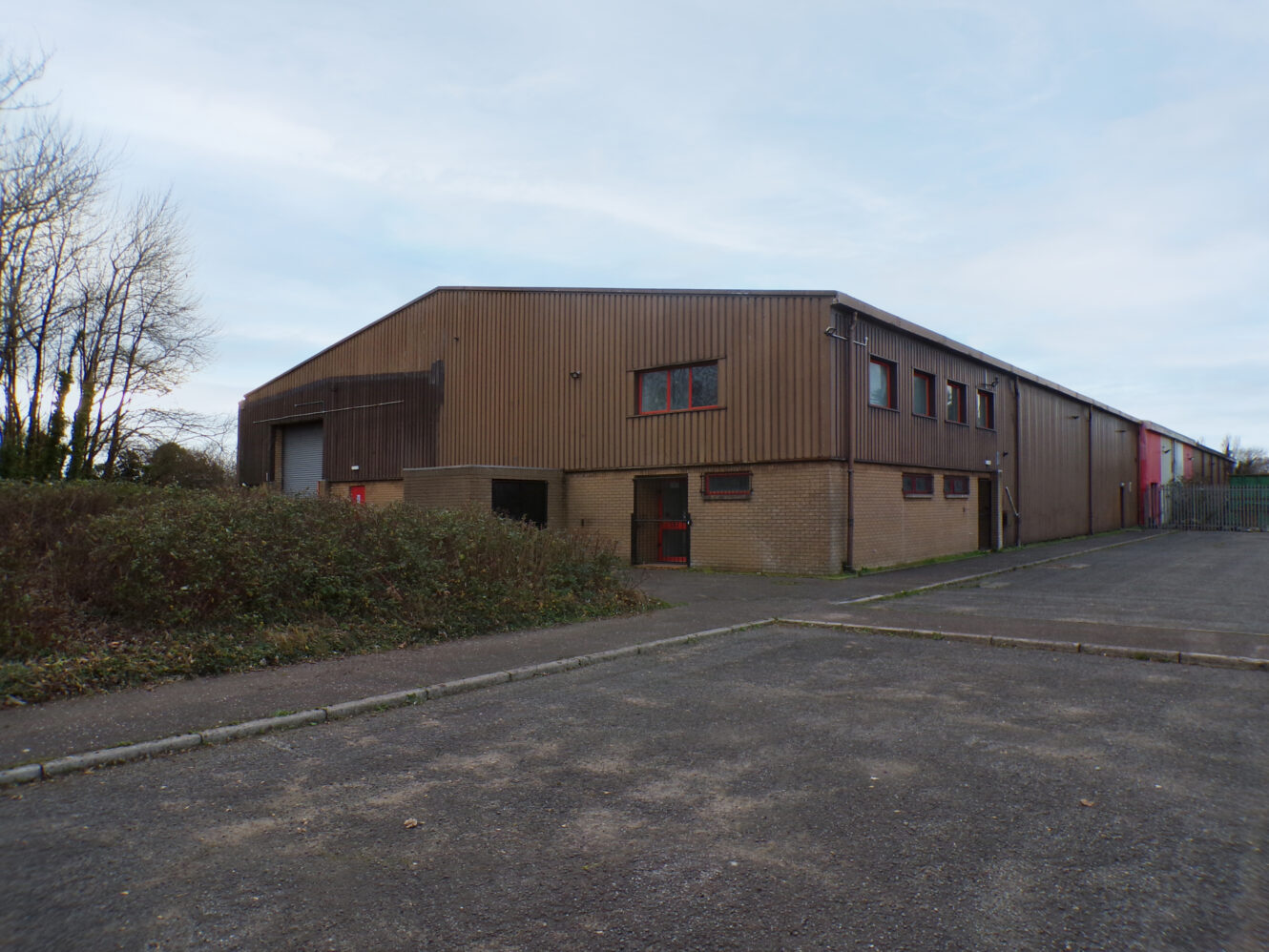 A large, rectangular industrial warehouse with brown metal siding, red doors, and a concrete parking lot in front. Trees and bushes are visible along the side.