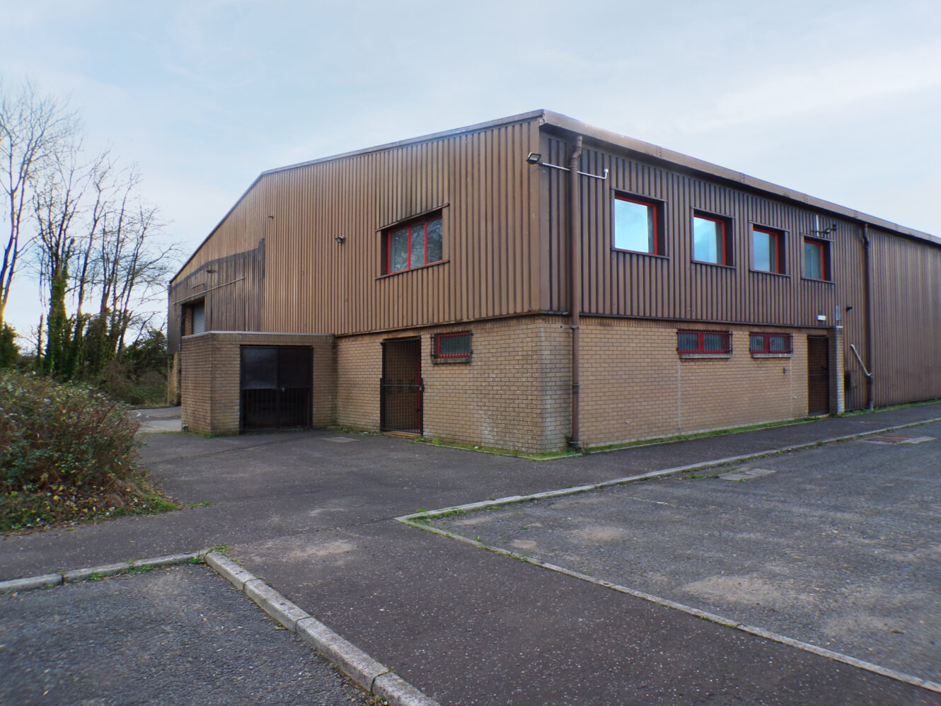 A large, brown industrial building with red-framed windows, surrounded by an empty parking lot and some sparse trees in the background.