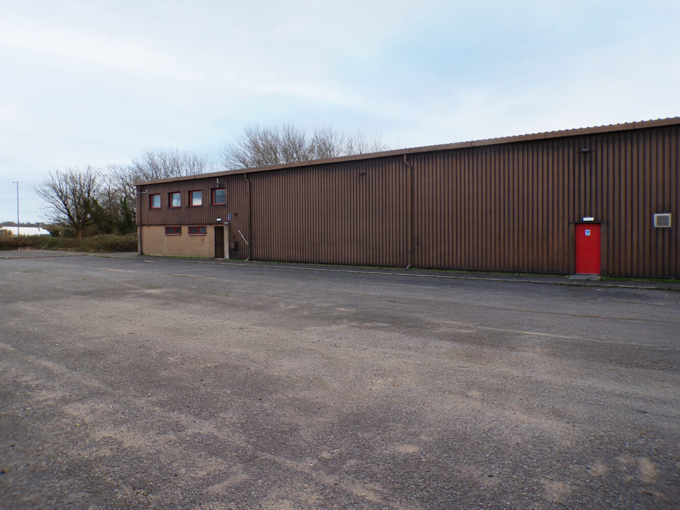 A large, brown industrial building with a red door and small windows, set on an empty paved lot with a few leafless trees in the background.