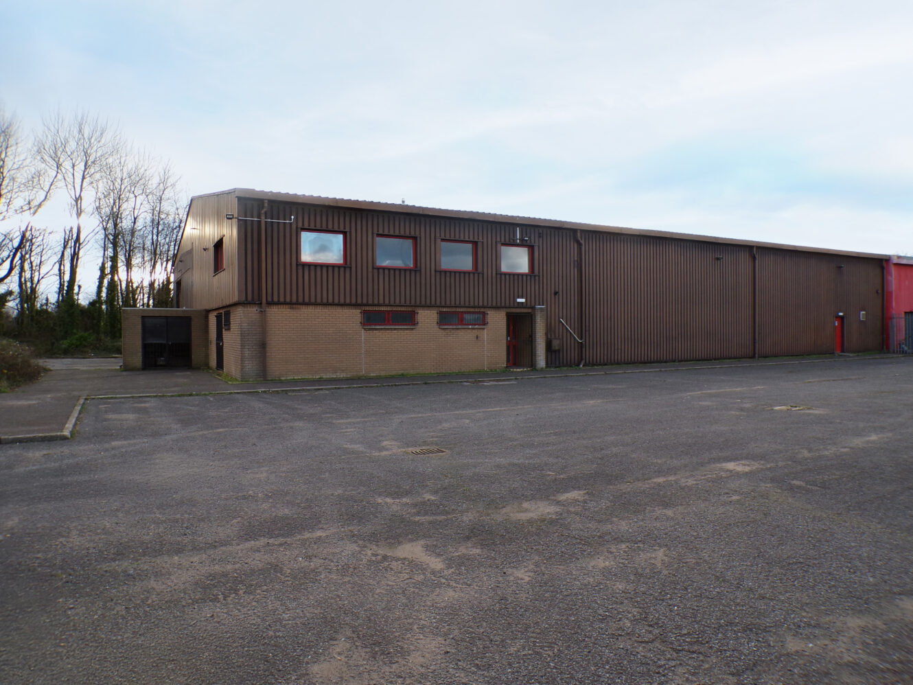 A large, rectangular industrial building with brown metal siding and small windows, situated next to an empty paved lot and surrounded by leafless trees.