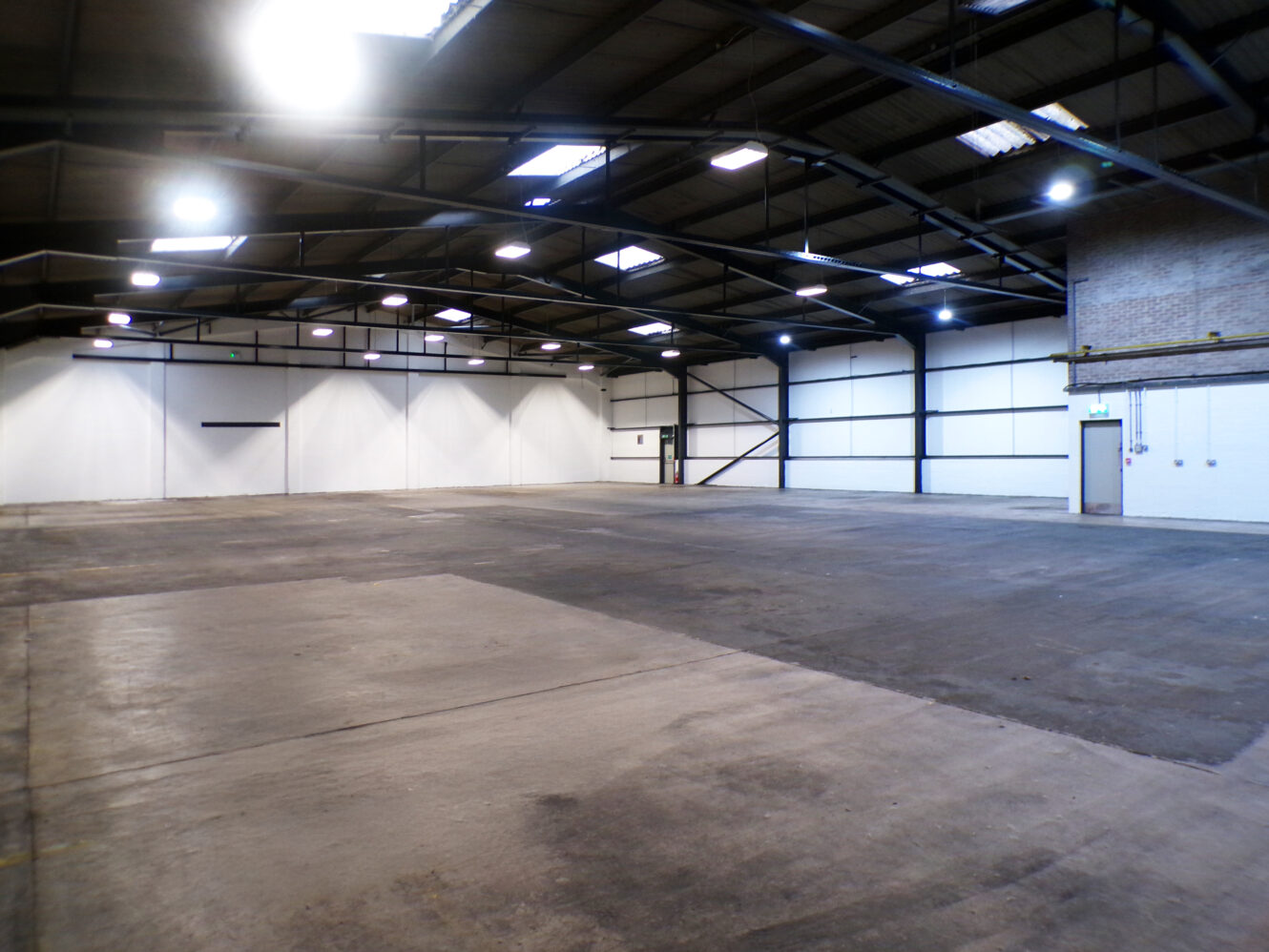 Large empty warehouse interior with concrete floor, high ceiling, white walls, overhead lights, and exposed beams. A closed door and electrical panels are visible on the right side.