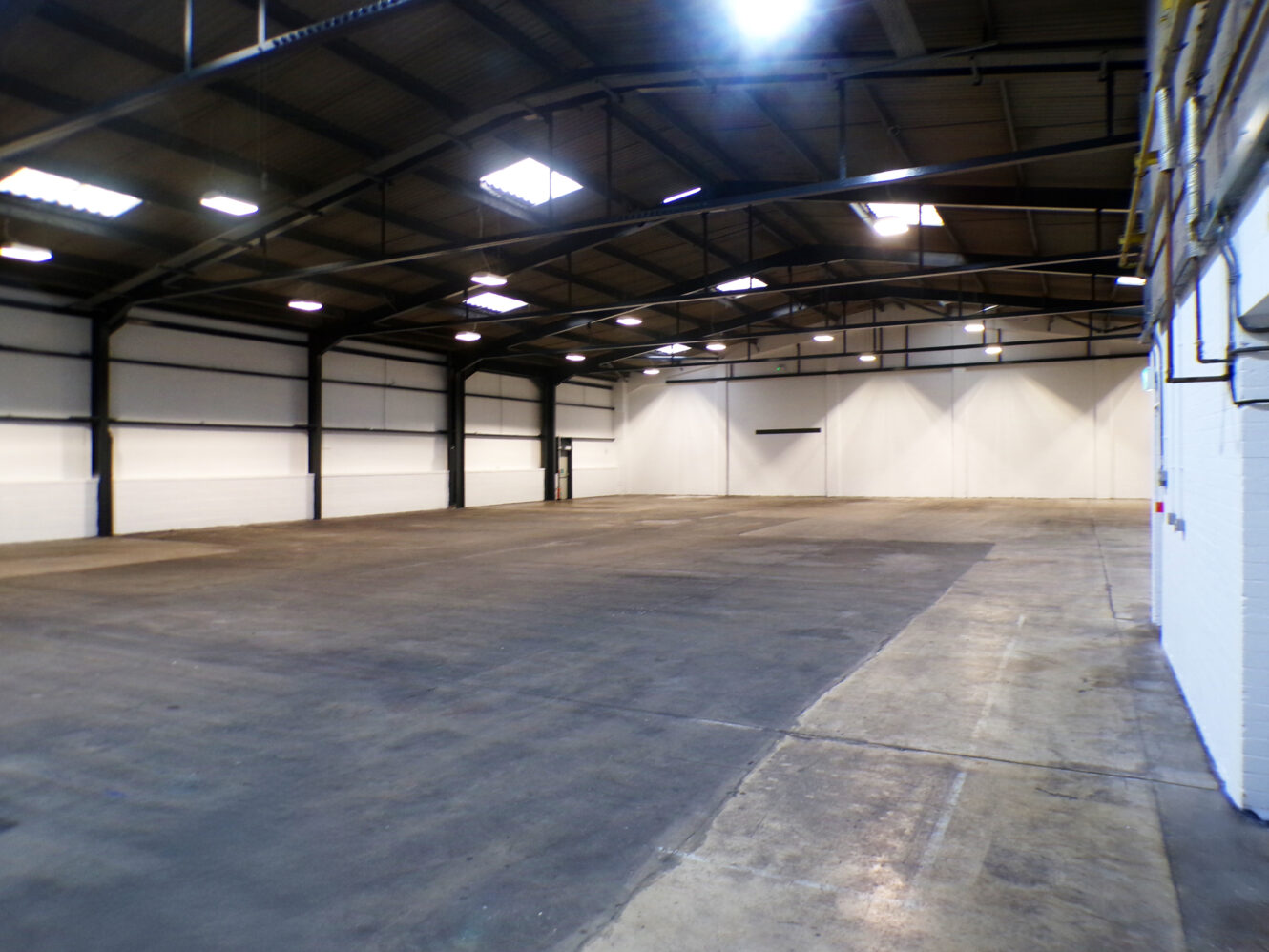 Spacious empty warehouse interior with high ceilings, exposed beams, white walls, and concrete flooring under bright ceiling lights.
