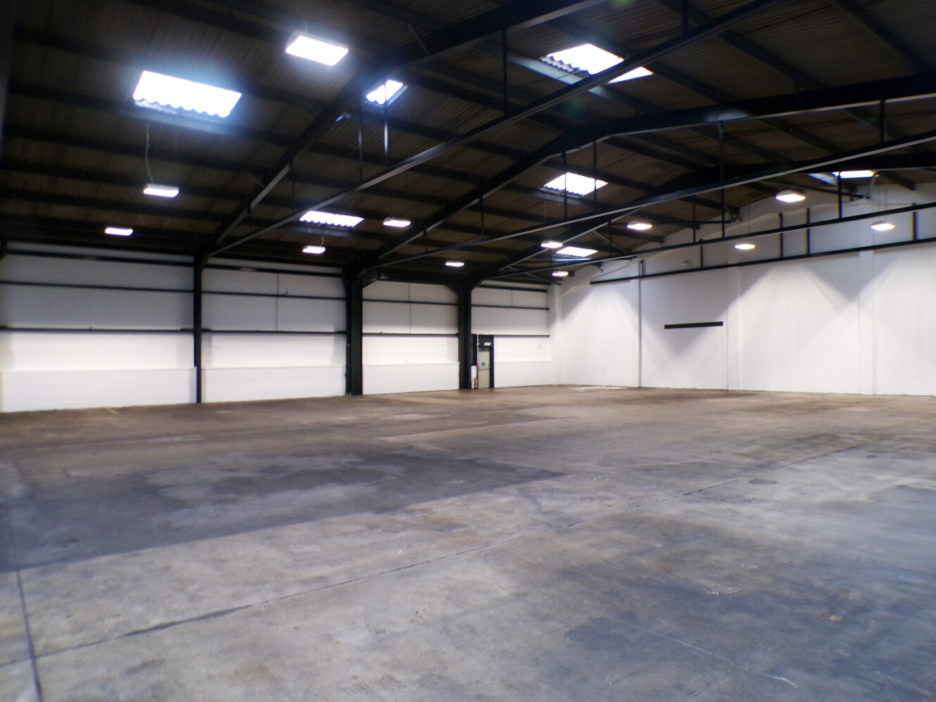 Large empty industrial warehouse with concrete floor, white walls, and exposed metal beams under a high ceiling with overhead lighting.