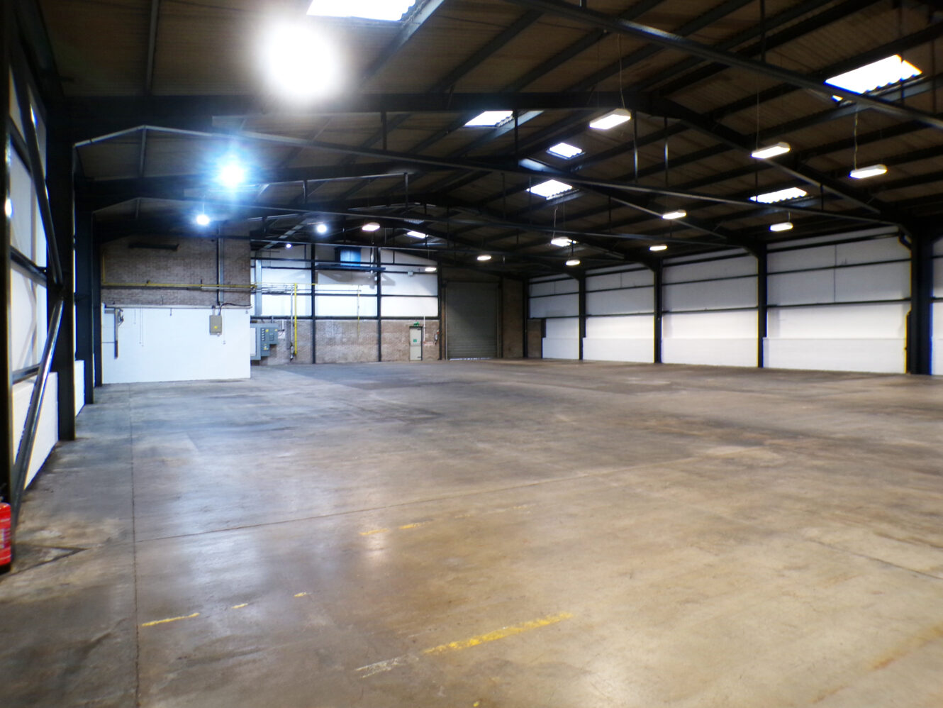 Large, empty warehouse with concrete floors, high ceilings, exposed beams, and bright overhead lighting. White walls and a small utility area are visible in the background.