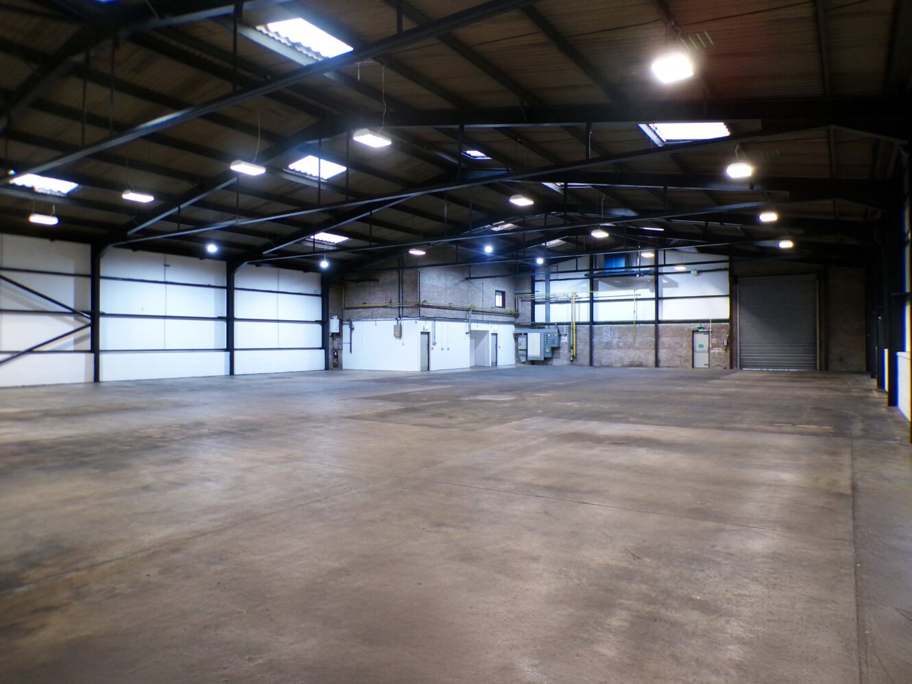 Spacious, empty warehouse interior with concrete floor, high ceiling, exposed beams, white walls, and industrial lighting.