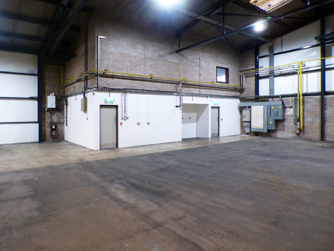 Interior of an empty industrial warehouse with concrete floors, exposed brick walls, overhead piping, control panels, and fluorescent lighting.