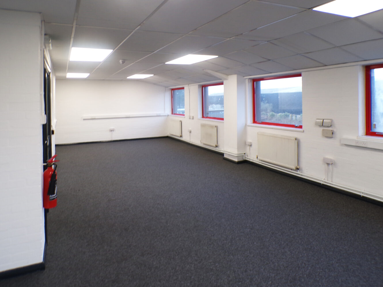 Empty office space with white walls, gray carpet, red-trimmed windows, ceiling lights, and a fire extinguisher mounted on the wall.