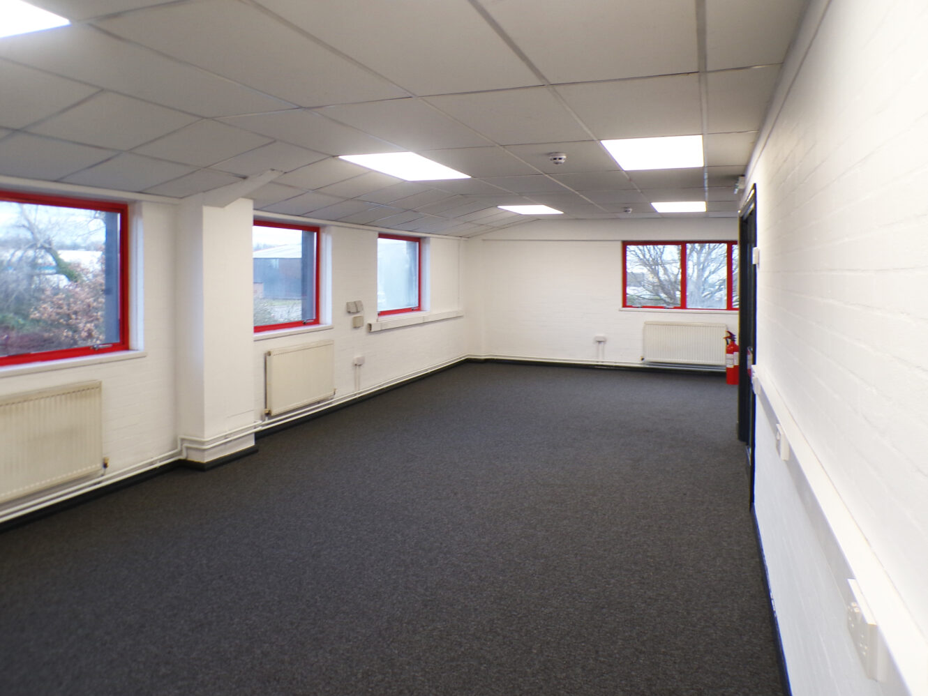 Empty office room with white walls, grey carpet, red-trimmed windows, fluorescent ceiling lights, and radiators along the walls.