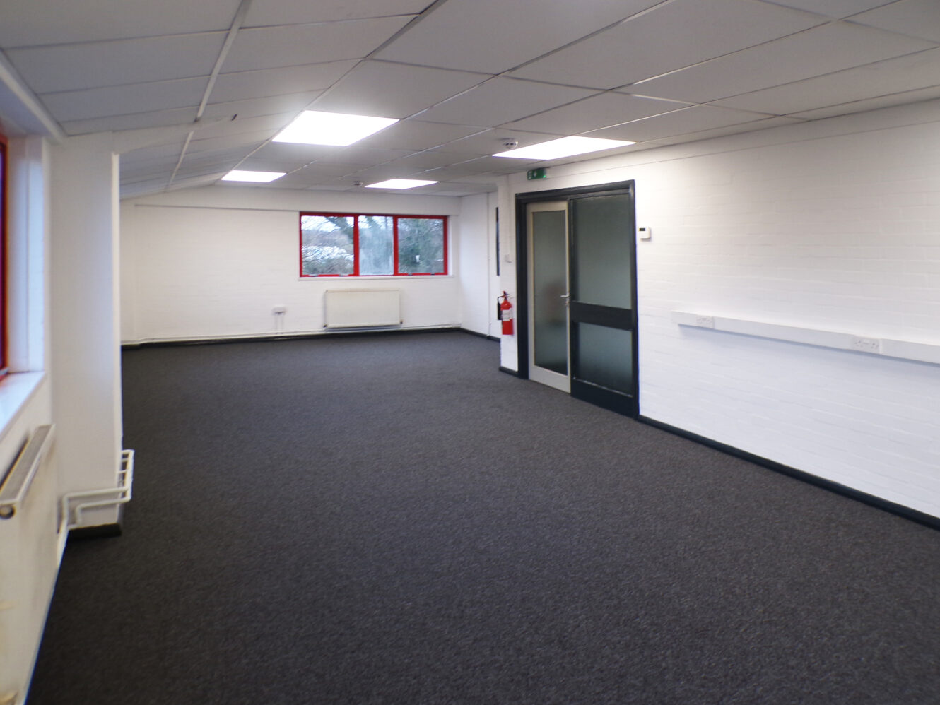 Empty office room with white walls, dark carpet, ceiling lights, red-framed windows, a radiator, a glass door, and a fire extinguisher mounted on the wall.