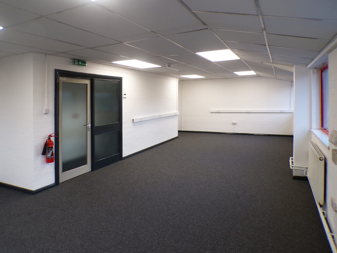 Empty office space with white walls, dark carpet, fluorescent ceiling lights, black-framed door, red window frame, and a fire extinguisher mounted near the entrance.
