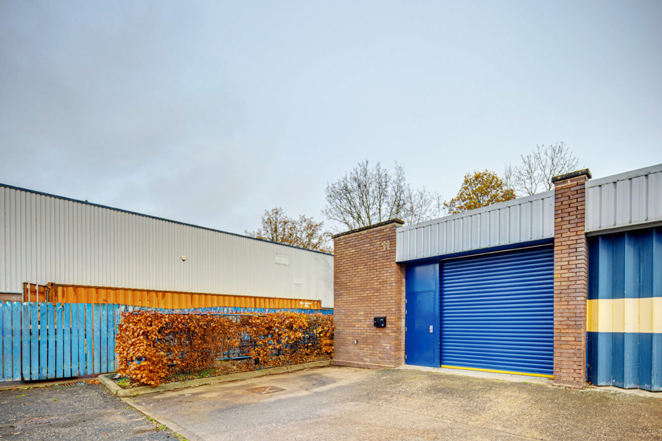Industrial building with a blue roller shutter door, brick wall, and a small parking area in front. Bushes and a blue wooden fence are visible to the left. Sky is overcast.
