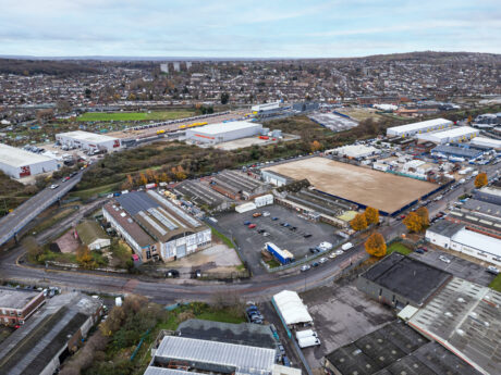 Aerial view of an industrial estate with warehouses, roads, parked vehicles, and residential areas in the background under a cloudy sky.