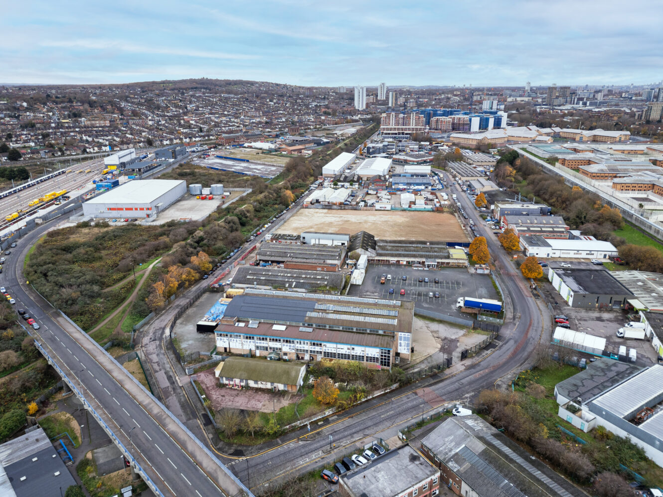 Aerial view of an industrial area with warehouses, roads, sparse trees, and a cityscape in the background under a cloudy sky.