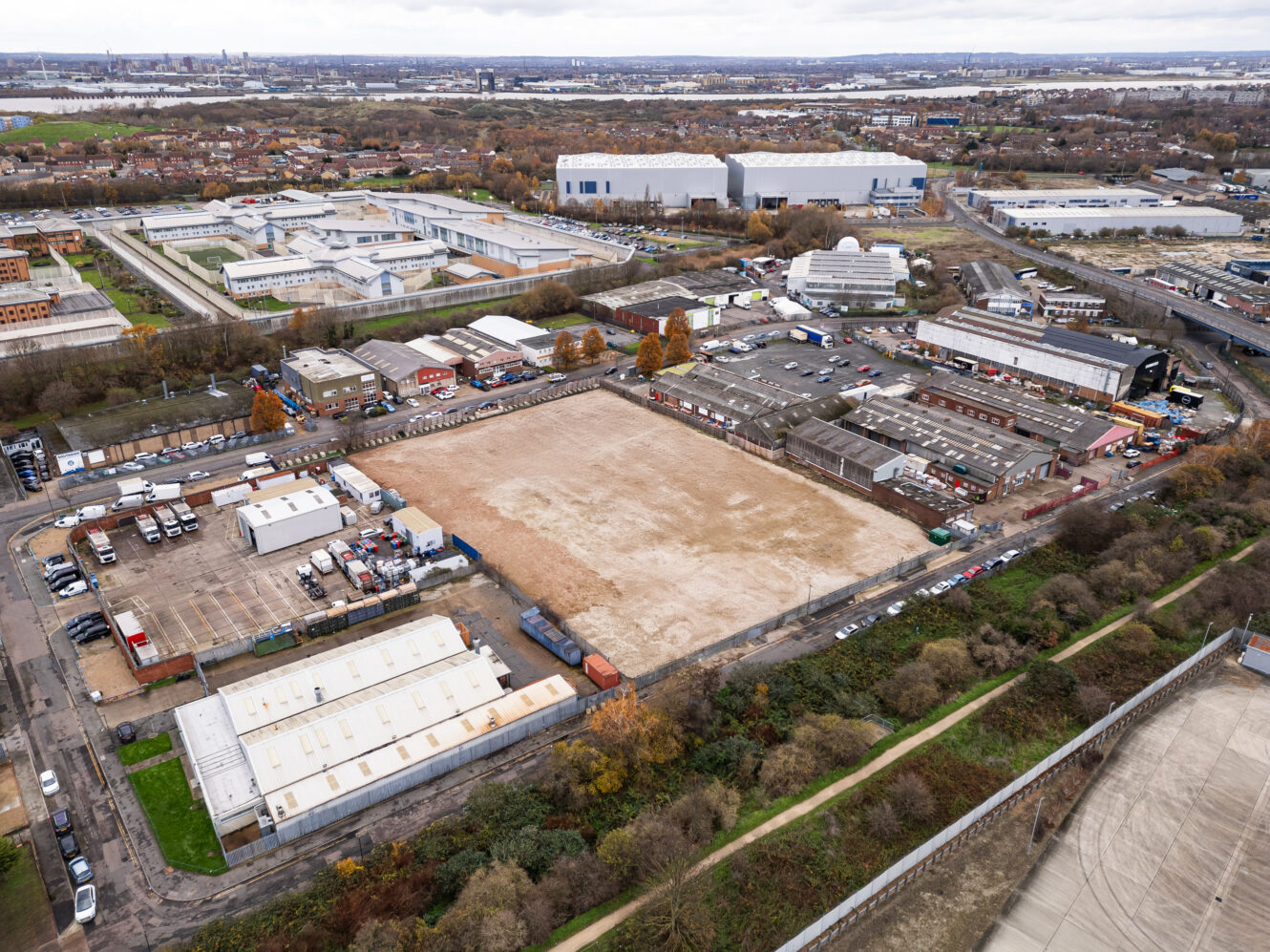 Aerial view of an industrial area with warehouses, parked vehicles, and a large empty lot surrounded by buildings and greenery.