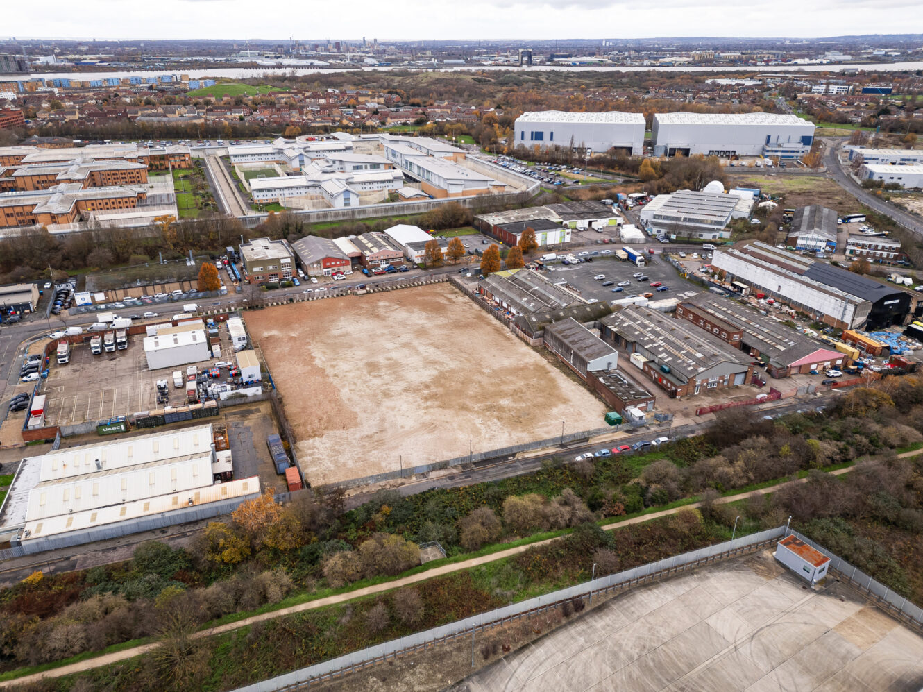 Aerial view of an industrial area with warehouses, parking lots, and a large rectangular dirt plot in the center, surrounded by roads and sparse vegetation.